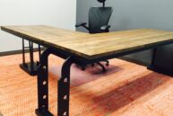 A modern office setup with a large L-shaped wooden desk on a red patterned rug. The desk has black metal supports and an ergonomic black chair with a headrest is placed behind it. The background wall is gray.