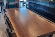 A large, rustic wooden table with a natural edge, surrounded by dark high-back chairs in a modern dining area. Shelves with bottles line the wall, and large windows in the background let in natural light.