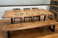 A wooden dining table set with six intricately designed wicker placemats, accompanied by a matching bench and three wooden chairs, all set on a polished hardwood floor. A shelf with glassware is visible on the right.
