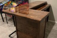 A polished wooden bar with a corner design stands on a carpeted floor, adjacent to vintage pinball machines with colorful designs. A metal footrest is attached to the bottom of the bar.