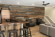 Spacious room with a rustic bar, wooden stools, wood-paneled accent wall, and light wood flooring. Includes a gray sectional sofa, dark chairs, and a modern ceiling fan. Stairs leading upwards are visible in the background.