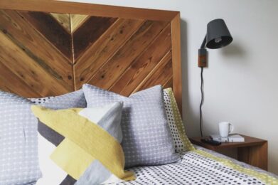 A cozy bedroom with a wooden headboard, geometric-patterned bedding in gray and yellow tones, and a matching pillow. A black wall-mounted lamp is above a wooden nightstand, which holds a white mug and a small notepad with a pen.