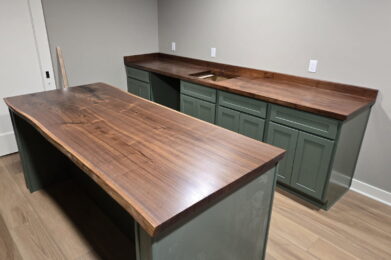 A kitchen with green cabinets and smooth, dark wooden countertops. An island with matching wood top and green base sits in the center. The room has light-colored walls and hardwood flooring.