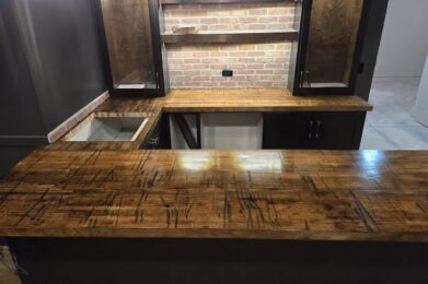 A home bar with a rustic wooden countertop and brick backsplash. Cabinets with glass doors sit above the counter. The area is dimly lit by hanging lights, creating a cozy atmosphere.