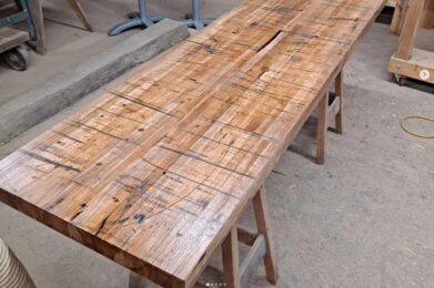 A long wooden table with a rustic, textured surface is set in a woodworking shop. Various tools and pieces of wood are in the background, placed on shelves and workbenches. The space has an industrial, handcrafted feel.