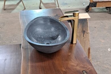 A live-edge wooden countertop with a smooth black stone vessel sink and a sleek gold faucet. The countertop showcases natural wood grain and texture, creating a rustic yet modern aesthetic.