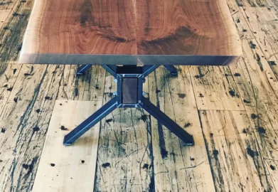 A Live Edge Tops wooden table with a natural edge design rests on a rustic wooden floor. The table showcases visible wood grain and is supported by metal legs, while the weathered look of the floor enhances its natural aesthetic.
