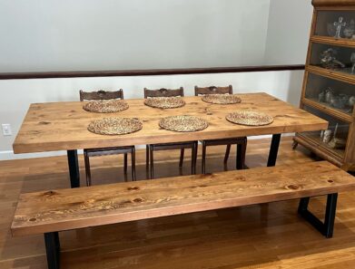 A wooden dining table set with six intricately designed wicker placemats, accompanied by a matching bench and three wooden chairs, all set on a polished hardwood floor. A shelf with glassware is visible on the right.