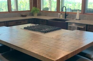 A spacious kitchen with a large wooden island featuring a built-in stovetop. There are cushioned bar stools around the island. Large windows provide a view of trees and greenery outside, creating a bright and inviting space.