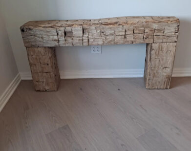 Rough-hewn wooden mantelpiece with a natural, rustic look, placed against a plain white wall on light wood flooring. An electrical outlet is visible on the wall behind it.
