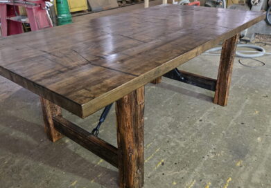 A large, rustic wooden table with thick legs and a patterned top sits in a workshop surrounded by tools, supplies, and industrial equipment.