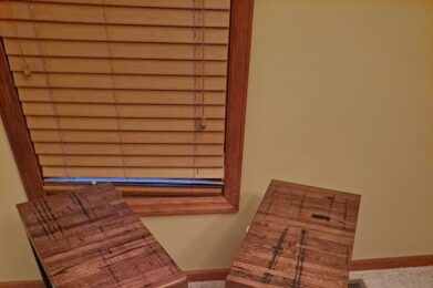 Two wooden side tables with dark metal legs are placed on a beige carpet. They are positioned at an angle, with a window with wooden blinds in the background, set against a light-colored wall.