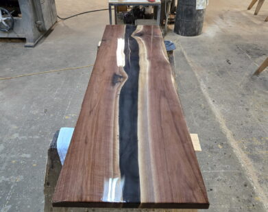 A long wooden table with a glossy finish and a dark, wavy resin river running through its center sits on sawhorses in a workshop filled with tools and wood materials.