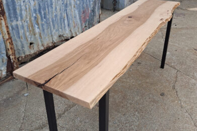 A long wooden table with a natural live edge sits on a concrete surface. The table has black metal legs and is set against a backdrop of weathered metal walls and a door. The wood grain is visible, showcasing its raw beauty.