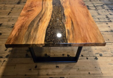A polished wooden table with a black resin river running through the center stands on a wooden floor, with samples of wood panels displayed on the wall behind it.