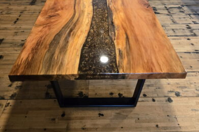 A polished wooden table with a black resin river running through the center stands on a wooden floor, with samples of wood panels displayed on the wall behind it.