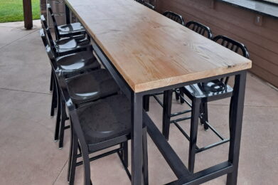 Outdoor covered patio with a long wooden dining table and black chairs. A silver grill is at the end of the table. The patio overlooks a scenic yard with green grass, trees, and a white fence under a cloudy sky.