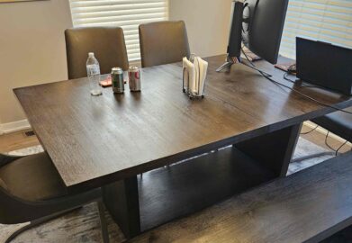 A dark wooden dining table with a matching bench and chairs, set with drinks, a water bottle, napkins, and a computer monitor, in a room with light-colored walls and windows with blinds.