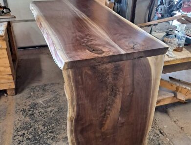 A polished wooden bar with a natural edge design is shown in a workshop setting. The wood has a rich, dark grain and is supported by a sturdy base. Workshop tools and materials are visible in the background.