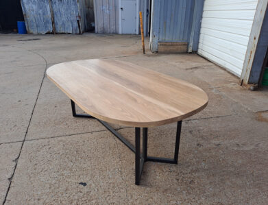 A wooden table with a smooth, oval-shaped top and metal legs is placed on a concrete surface outside a building. The background features industrial elements like corrugated metal walls and a partially open garage door.