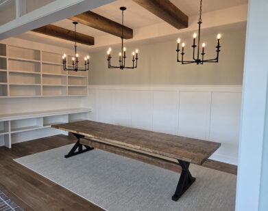 A spacious room featuring a rustic wooden table on a light rug, open white built-in shelves along the wall, two black metal chandeliers, wood ceiling beams, and white paneled walls.