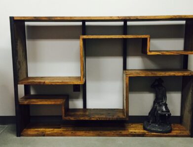A wooden, geometrically designed bookcase with staggered shelves stands against a white wall. A small black statue of a cowboy is placed on the bottom right shelf, adding a decorative touch to the minimalist furniture piece.