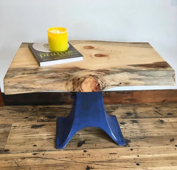 A small wooden table with a blue metal base sits on a wooden floor. A yellow candle and a book titled prattle rest on the tables surface, which features natural wood knots and grain patterns.