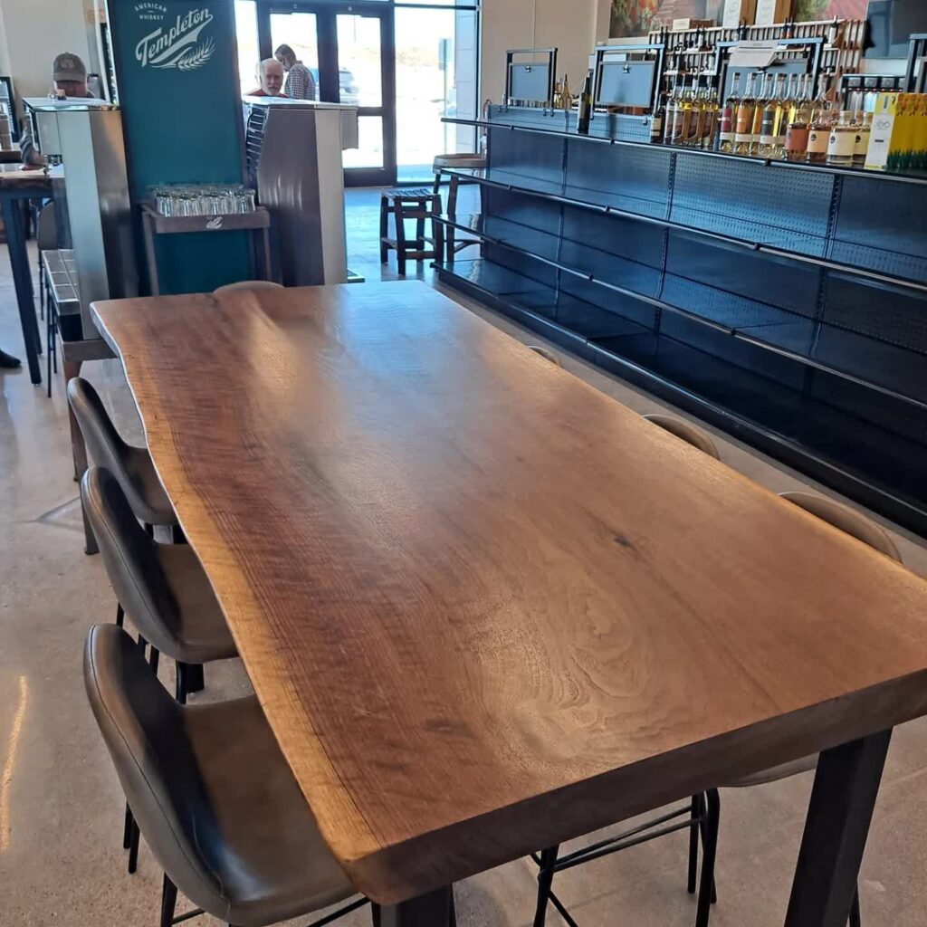 A large, rustic wooden table with a natural edge, surrounded by dark high-back chairs in a modern dining area. Shelves with bottles line the wall, and large windows in the background let in natural light.