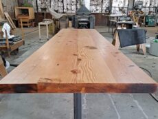 A large wooden table with a natural finish is set in a workshop. The table rests on a sturdy black metal frame. The workshop background includes various woodworking tools, machines, and materials.