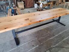 A long, rustic wooden table with a sturdy black metal frame standing on a concrete floor in a workshop. Various tools and materials are visible in the background.