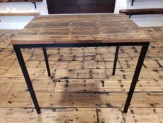 A rustic wooden table with a metal frame is positioned on a wooden plank floor. The tables surface shows natural wood grain patterns and knots, complementing the surrounding wooden decor.
