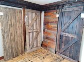 A display of four rustic wooden doors in a showroom, each with different textures and shades of brown. The doors feature styles such as barn and plank, and the floor is made of matching wood panels.