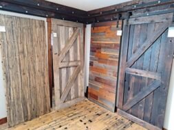 A display of four rustic wooden doors in a showroom, each with different textures and shades of brown. The doors feature styles such as barn and plank, and the floor is made of matching wood panels.