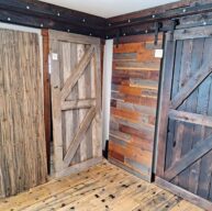 A display of four rustic wooden doors in a showroom, each with different textures and shades of brown. The doors feature styles such as barn and plank, and the floor is made of matching wood panels.