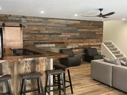 Spacious room with a rustic bar, wooden stools, wood-paneled accent wall, and light wood flooring. Includes a gray sectional sofa, dark chairs, and a modern ceiling fan. Stairs leading upwards are visible in the background.