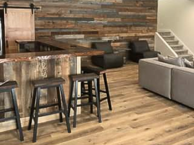 Spacious room with a rustic bar, wooden stools, wood-paneled accent wall, and light wood flooring. Includes a gray sectional sofa, dark chairs, and a modern ceiling fan. Stairs leading upwards are visible in the background.