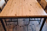 A wooden table with a polished surface stands on a rustic wooden floor. Surrounded by four matching chairs, the setting is in a well-lit room with additional tables in the background.