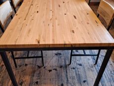 A wooden table with a polished surface stands on a rustic wooden floor. Surrounded by four matching chairs, the setting is in a well-lit room with additional tables in the background.