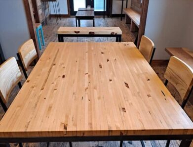 A wooden table with a polished surface stands on a rustic wooden floor. Surrounded by four matching chairs, the setting is in a well-lit room with additional tables in the background.