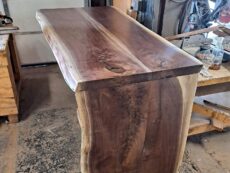 A polished wooden bar with a natural edge design is shown in a workshop setting. The wood has a rich, dark grain and is supported by a sturdy base. Workshop tools and materials are visible in the background.