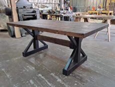 A sturdy wooden table with a dark, rustic finish and black metal X-shaped legs is positioned in a workshop. Various tools and equipment can be seen in the background, creating an industrial setting.
