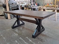 A sturdy wooden table with a dark, rustic finish and black metal X-shaped legs is positioned in a workshop. Various tools and equipment can be seen in the background, creating an industrial setting.