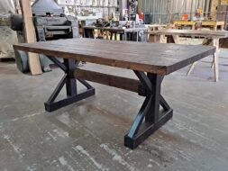 A sturdy wooden table with a dark, rustic finish and black metal X-shaped legs is positioned in a workshop. Various tools and equipment can be seen in the background, creating an industrial setting.
