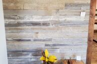 A rustic wooden wall with a small black vent at the top. Below, a colorful houseplant in a white pot stands next to a small wooden table with turquoise legs. The floor is made of reclaimed wood.