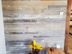 A rustic wooden wall with a small black vent at the top. Below, a colorful houseplant in a white pot stands next to a small wooden table with turquoise legs. The floor is made of reclaimed wood.
