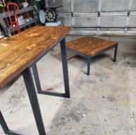 Wooden tables with metal legs on a concrete garage floor. The larger table is to the left, with a square table to the right. The garage in the background shows various tools and equipment.