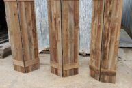 Three rustic wooden panels stand upright on a concrete surface in front of a corrugated metal wall. Each panel is made of vertical planks with horizontal supports at the top and bottom. The wood appears weathered and textured.