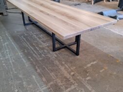 A long wooden table with a natural finish and black metal legs is set in a workshop. The surrounding space has woodworking tools and materials, visible in the background.