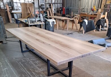 A long wooden table with a natural finish and black metal legs is set in a workshop. The surrounding space has woodworking tools and materials, visible in the background.