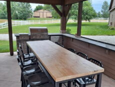 Outdoor covered patio with a long wooden dining table and black chairs. A silver grill is at the end of the table. The patio overlooks a scenic yard with green grass, trees, and a white fence under a cloudy sky.